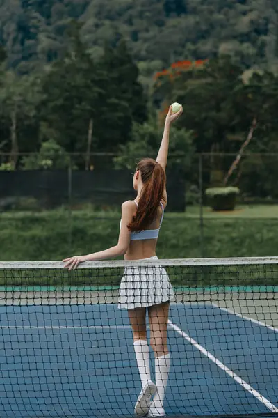 Enerjik genç bayan tenis oyuncusu güneşli bir sahada topa servis yapıyor. Çevresi yemyeşil, odak ve atletizmle çevrili.