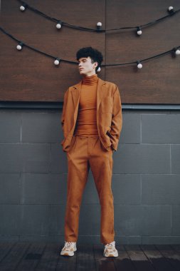 Kahverengi balıkçı yaka ve takım elbise giyen şık bir erkek model, gri fayanslı bir zeminde duruyor, sıcak ışıklandırmayla çevrili.