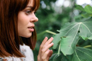 Dalgın bir kadın ağaçta canlı yeşil bir yaprağa, düşünceli bir duruşla el üstünde bakar.