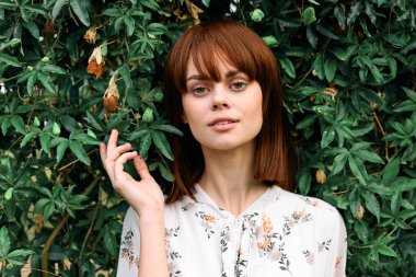 Canlı kızıl saçlı kadın gür yeşil çalıların önünde yapraklarla poz veriyor.