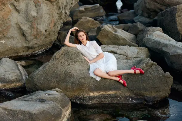 Beyaz elbiseli sakin bir kadın suda kayanın üzerinde oturuyor, kırmızı ayakkabılar dikizliyor.