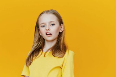 Uzun saçlı, parlak sarı tişörtlü, kendine güvenen genç bir kız, neşeli sarı bir arka plana karşı duruyor, gençlik modası ve pozitifliği için ideal zevk ve stil sunuyor.