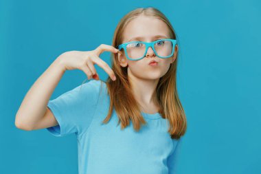 Gözlüklü genç bir kız, gençliği ve modern stili temsil eden parlak mavi bir zemin üzerinde barış işaretiyle rahatça poz veriyor. Kendini ifade etme ve eğlenme kavramları için mükemmel.