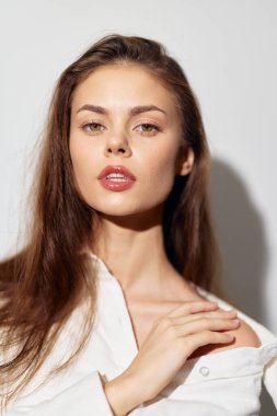 Güzel bir genç kadının yakın portresi, uzun saçları ve yumuşak yüz hatlarıyla rahat bir duruşu olan beyaz bir gömlek giyiyor, özgüven ve zarafet yayıyor.
