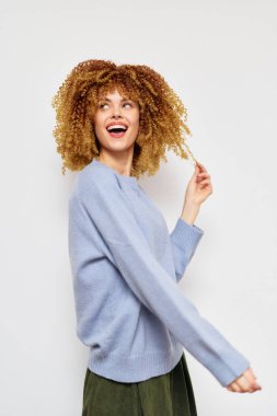 Açık mavi kazak giymiş neşeli kıvırcık saçlı genç bir kadın, basit bir beyaz arkaplana karşı duruyor. Neşeli ve özenli bir saç teli tutuyor.