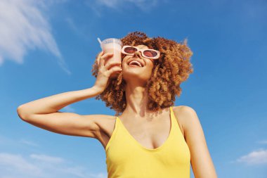 Kıvırcık saçlı neşeli bir kadın, parlak bir arkaplanda şık güneş gözlüğü ve sarı bir mayo takıyor.