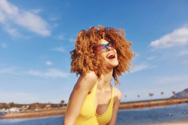 Kıvırcık saçlı, güneş gözlüğü takan neşeli genç bir kadın kumsalda gülümsüyor, açık mavi bir gökyüzü ve arka planda su var.