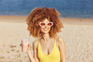 Kıvırcık saçlı, güneş gözlüğü takan ve sarı mayo giyen neşeli kadın deniz kenarında serinletici bir içkinin keyfini çıkarıyor.
