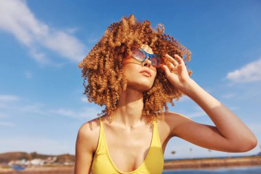 Kıvırcık saçlı, sarı mayo giymiş, açık mavi gökyüzü ve yumuşak bulutlarla güneşli bir sahilde dinlenen neşeli bir kadının canlı bir sahnesi.