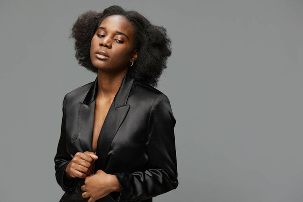 Portrait of a confident young woman wearing a chic black satin blazer, showcasing her natural curly hair and poised expression in a minimalistic gray setting
