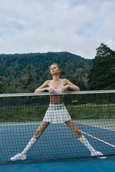 Beyaz spor giyimli dinamik genç bayan tenisçi, zengin dağlarla çevrili tenis kortunda poz verirken kendine güveni tam, spor ve spor tutkusu yansıtıyor.