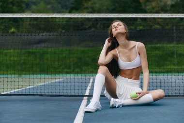 Spor elbiseli genç bir kadın, elinde yeşil bir tenis topuyla tenis kortunda oturuyor. Yüzü rahatlamış ve odaklanmış, spor, zindelik ve spor temalarını somutlaştırıyor.