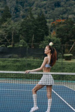 Açık mavi bir spor bluzu ve beyaz bir etek giyen kendinden emin bir bayan tenisçi, tenis kortunda poz veriyor, canlı bir açık hava ortamında azim ve atletizmi somutlaştırıyor.