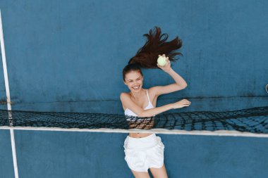 Spor kıyafeti giymiş, mutlu bir Asyalı kadının oynadığı enerjik bir spor sahnesi dinamik mavi arka plan ve parlak tenis topu canlı atmosferi güçlendiriyor.