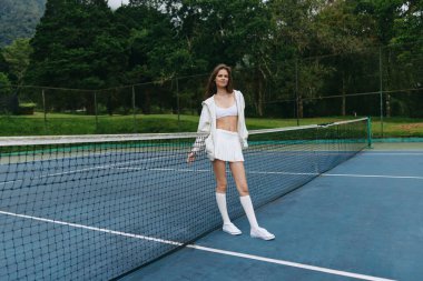 Beyaz spor kıyafetleri giymiş kendinden emin genç bir bayan tenisçi bir tenis kortunda duruyor, coşku ve neşe saçıyor yemyeşil arka plan canlı açık hava atmosferini arttırıyor, mükemmel bir ortam için.