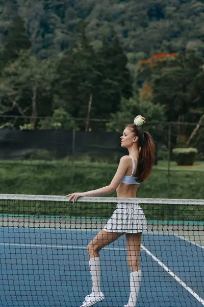 Açık mavi bir spor bluzu ve beyaz bir etek giyen kendinden emin bir bayan tenisçi, tenis kortunda poz veriyor, canlı bir açık hava ortamında azim ve atletizmi somutlaştırıyor.