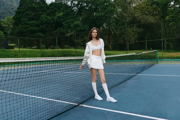 Beyaz spor kıyafetleri giymiş kendinden emin genç bir bayan tenisçi bir tenis kortunda duruyor, coşku ve neşe saçıyor yemyeşil arka plan canlı açık hava atmosferini arttırıyor, mükemmel bir ortam için.