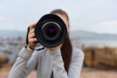 Sahilde duran genç bir kadın, elinde kamerayla izleyiciye bakıyor, doğal bir kıyı ortamında fotoğrafçılığa olan hevesini dile getiriyor.