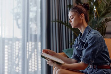 Kanepede uzanan ve rahat ev ortamında kot gömlekle kitap okuyan bir kadın.