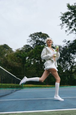 Beyaz spor elbiseli enerjik genç bir kadın tenis kortunda mutlu bir şekilde koşuyor, bir içki tutuyor, yemyeşil bir ortamda sağlıklı ve aktif bir yaşam tarzını somutlaştırıyor.