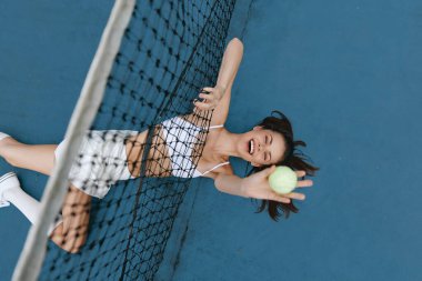 Genç bir kadının tenis kortunda eğlenmesi, yerde yatarken neşe göstermesi, aktif bir yaşam tarzı ve canlı renkler sergilemesi, spor temalı içerikler için ideal.