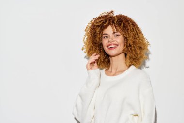 Kıvırcık saçlı neşeli bir kadın minimalist beyaz bir arka plana karşı duruyor, yumuşak bir kazak giyiyor neşeli ifadesi kendine güveni ve yaklaşılabilirliği temsil ediyor, yaşam tarzı için mükemmel.