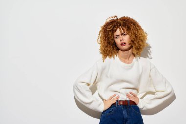 Moda mankeni, rahat beyaz bir kazakla moda kıvırcık saçlar sergiliyor, beyaz bir duvara karşı poz veriyor. Görüntü modern ve şık bir estetik, moda ve yaşam tarzı projeleri için mükemmel.