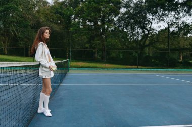 Spor kıyafetleri giymiş kendine güvenen genç bir kadın tenis kortunda duruyor, yemyeşil bir ortamda atletizm ve tarz aktarıyor, spor temalı içerikler için mükemmel.