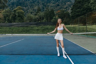 Spor kıyafetli atletik genç bir kadın tenis kortunda poz veriyor, spora olan güvenini ve bağlılığını gösteriyor. Canlı açık hava sahnesi onun sağlık ve zindeliğe olan bağlılığını vurguluyor.