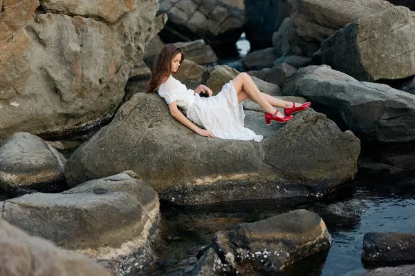 Okyanus kayasına oturmuş deniz manzarasına hayran beyaz elbiseli, dingin bir kadın.