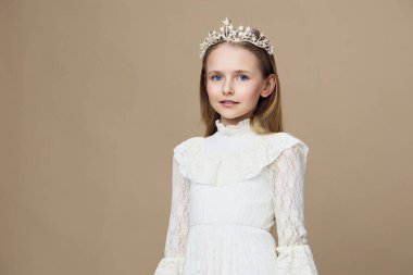 Beyaz dantelli bir elbise ve bir taç giymiş neşeli genç bir kızın portresi yumuşak bej bir arkaplanda duruyor, çocuk modası ve kutlama temaları için mükemmel.