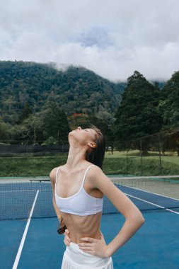 Tenis kortunda kendine güvenen genç bir bayan sporcu, beyaz bir spor kıyafeti giyiyor ve doğayı kucaklıyor. Huzurlu arka plan onun rahat ifadesini tamamlıyor.