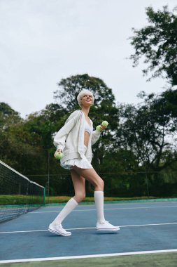 Spor kıyafetli neşeli genç bir kadın güneşli bir günde dışarıda tenis oynuyor, gülümsüyor ve tenis toplarını tutuyor, canlı yeşil bir geçmişi var, spor ve aktif yaşam temaları için mükemmel.