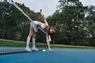 Tenis kıyafeti giymiş atletik bir kadın, topu yakalamak için ağdan eğilirken yeşil bir zemin yakalıyor ve sportif bir yaşam tarzını vurguluyor.