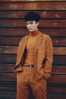 Kahverengi takım elbise ve balıkçı yaka giyen şık erkek manken, ahşap panellere karşı düşünceli bir şekilde poz veriyor.