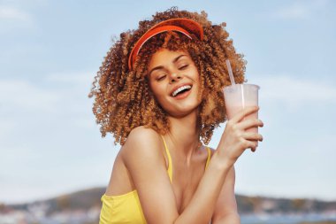 Kıvırcık saçlı neşeli bir kadın güneşli bir zemine kurulmuş güneşlik takan ferahlatıcı bir içecek tutar.