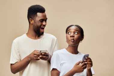 Neşeli bir siyah çift akıllı telefonlarıyla iletişim kuruyor, sürpriz ve meraklarını dile getiriyor, yumuşak bej bir arka plana kuruluyor, modern ilişkileri göstermek için ideal.