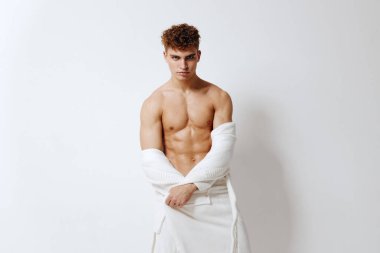 Güçlü erkek model, beyaz kumaşla örtülmüş, sade bir arka planda sergilenen atletik bir fiziğe sahip. Zindelik temaları için sağlıklı, güven ve gücü somutlaştırır..