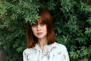 Doğada yemyeşil çalıların önünde poz veren kızıl saçlı canlı bir kadın.