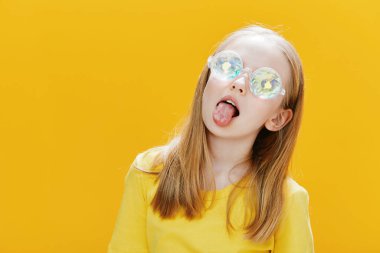 Zarif yansıtıcı gözlüklü neşeli genç bir kız ve sarı bir kıyafet, neşeli bir şekilde dilini parlak sarı bir zemin üzerine uzatıyor, mutluluğu ve gençliği somutlaştırıyor