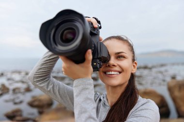 Spor kıyafetleri içinde neşeli bir genç kadın plajda büyüleyici doğa fotoğrafçılığını yakalıyor fotoğrafçılığa olan tutkusunu ve açık hava güzelliğini gözler önüne seriyor.