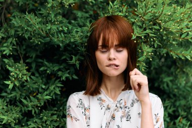 Canlı, kızıl saçlı bir kadın yemyeşil çalıların önünde dikilirken yüzüne dokunuyor.