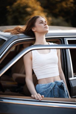 Beyaz bir bluz ve kot pantolon giymiş şık genç bir kadın, güneşli bir sonbahar gününde, güzel, sıcak bir arka planla kaygısız bir ruhu somutlaştıran, klasik bir arabadan dışarı sarkıyor..