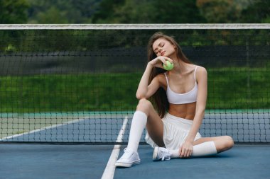Beyaz spor kıyafeti giymiş atletik bir kadın model tenis kortunda güven içinde oturuyor, bir tenis topu ile, yeşil bir zemine karşı enerji ve kararlılığı somutlaştırıyor.