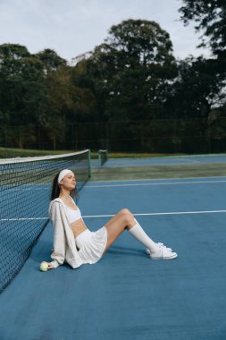 Genç bir bayan manken, spor kıyafetleri içinde mavi bir tenis kortunda dinleniyor, sükunet saçıyor, beyaz bir spor giysisi ve yeşil bir saç bandıyla sarılı, dingin bir sporu yansıtıyor.