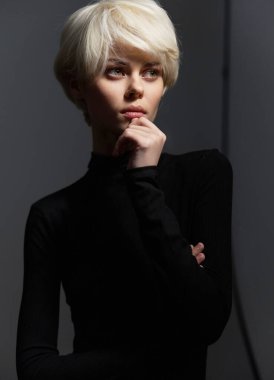 Minimalist siyah kıyafetli, platin sarısı saçlı dalgın bir kadın manken, yumuşak siyah bir arkaplanda duruyor, duygusal derinlik ve sanatsal ifade aktarıyor..