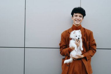 Kahverengi kıyafetli gülümseyen genç adam küçük beyaz bir köpekle poz veriyor, modern gri arka planda sıcak ve neşeli bir atmosfer yaratıyor.