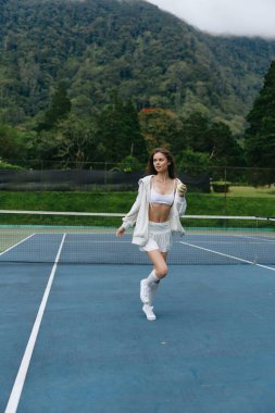 Enerjik genç bir kadın şık spor kıyafetleriyle tenis kortunda koşuyor, atletizmin ve canlılığın özünü yakalıyor canlı bir doğal zemine karşı.