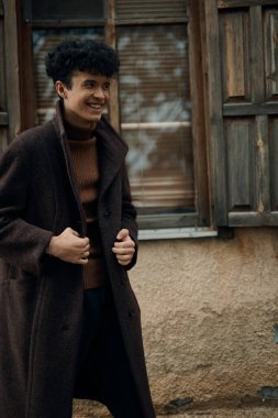 Siyah pantolon ve ayakkabılarla eski binanın önünde duran kahverengi ceketli ve kaplumbağa boyunlu moda adamı..