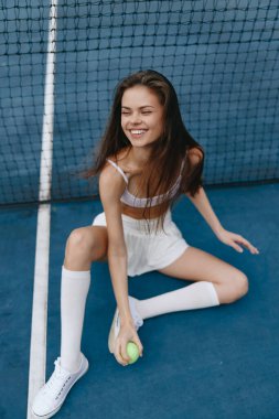 Spor kıyafetli neşeli genç bir kadın tenis kortunda oturuyor, elinde bir tenis topu tutarken sevinç ifade ediyor, spor ve spor kavramları için mükemmel.